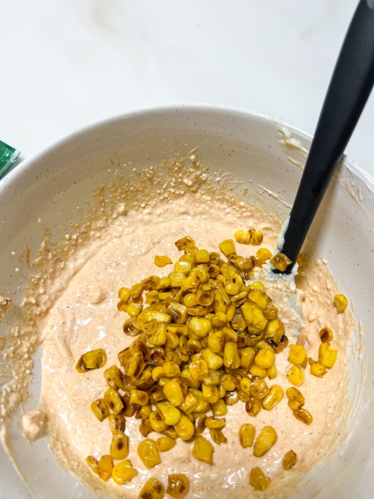 Charred corn kernels being added to a creamy spiced elote dip base in a white ceramic mixing bowl with a black spatula