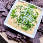 Elote dip in a white square bowl topped with cotija cheese and fresh cilantro, surrounded by blue corn tortilla chips on a round serving plate