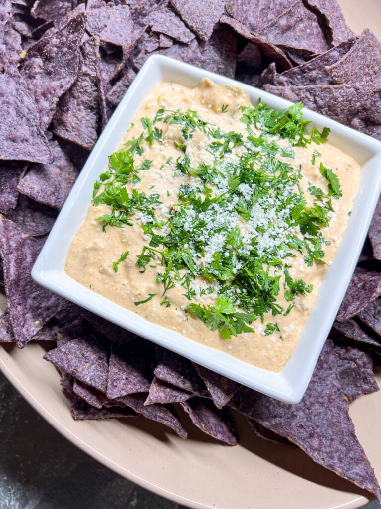Elote dip in a white square bowl topped with cotija cheese and fresh cilantro, surrounded by blue corn tortilla chips on a round serving plate