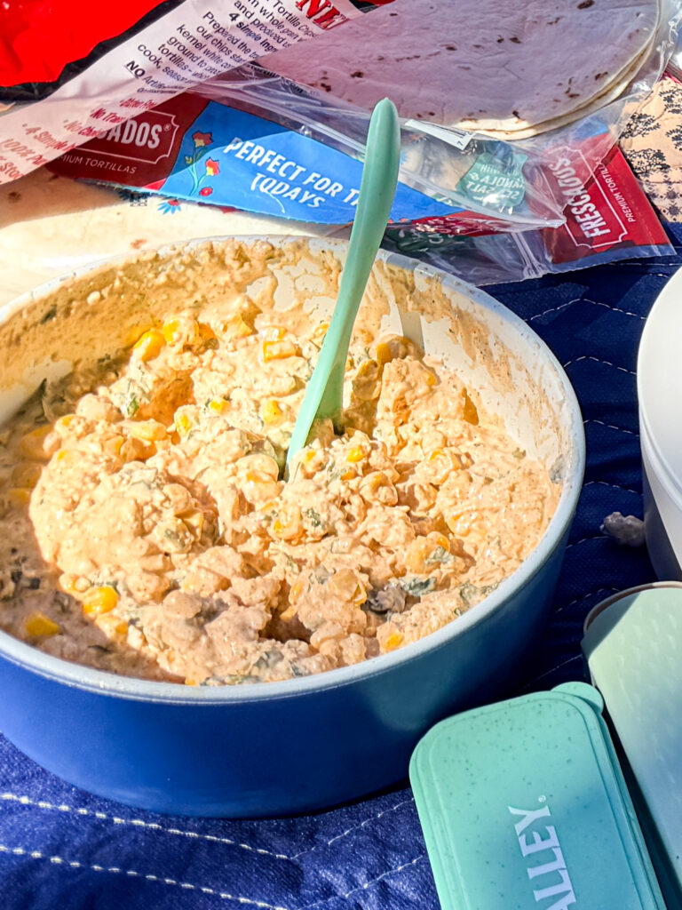 Elote dip in a blue ceramic bowl at an outdoor picnic table with tortilla packaging and chip bags in the background