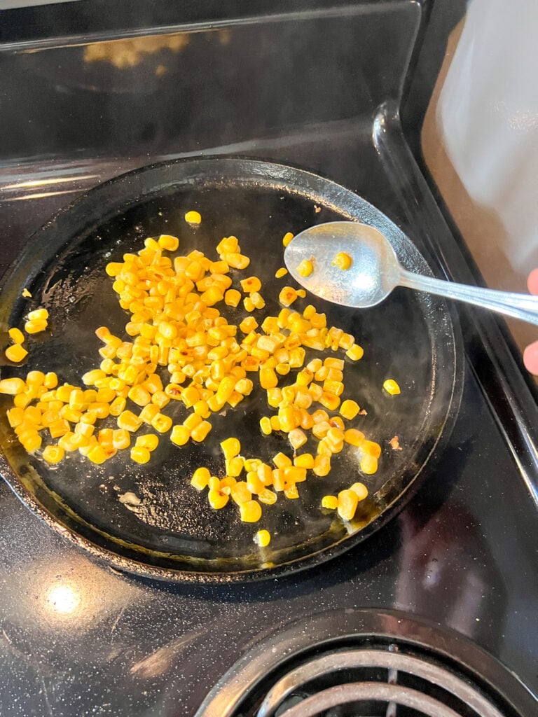 Canned corn kernels searing in a cast iron skillet on a stovetop, developing golden toasted color with a spoon resting nearby