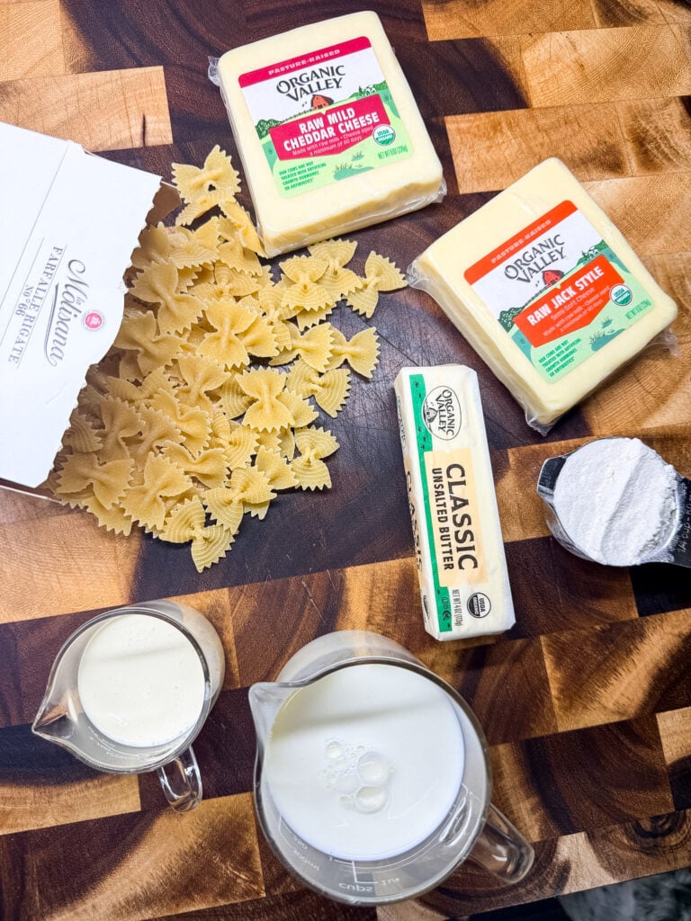 Mac and cheese ingredients flat lay on a wood cutting board — Organic Valley cheddar and jack cheese blocks, farfalle pasta, butter, milk, and heavy cream