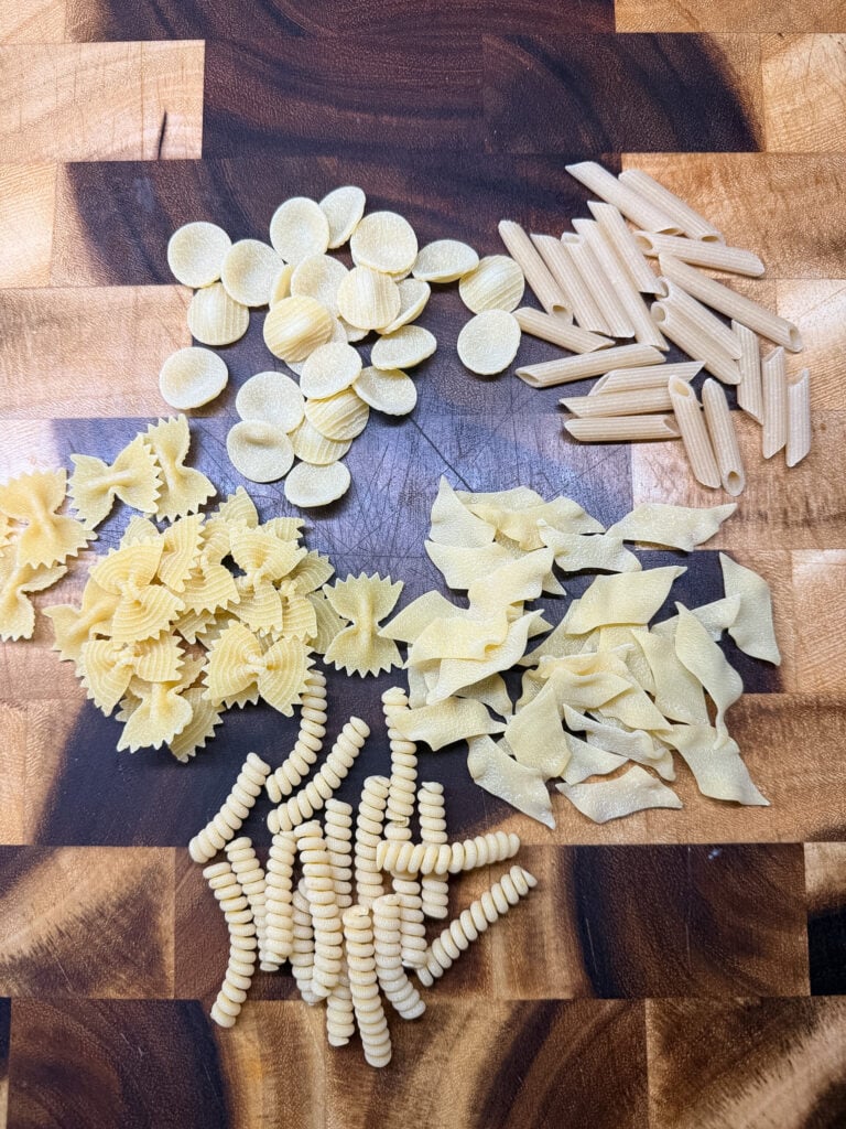 Variety of dry pasta shapes arranged on a wood cutting board — orecchiette, farfalle, penne, lasagne sheets, and cavatappi for mac and cheese