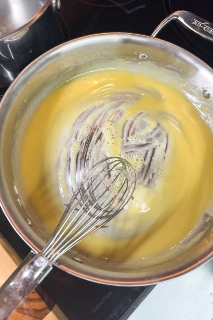 Golden roux beginning to thin as liquid is whisked in, whisk visible in All-Clad saucepan