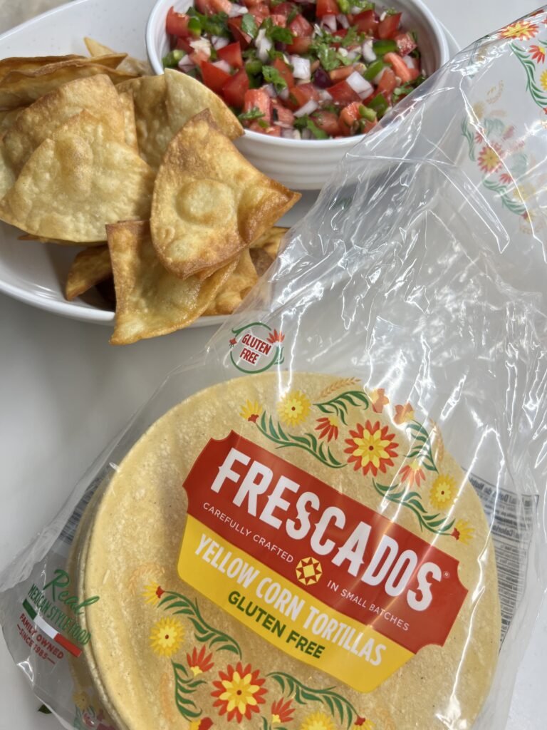 Crispy homemade tortilla chips piled on a white plate with a bowl of fresh diced tomato, cilantro, onion, and jalapeño pico de gallo