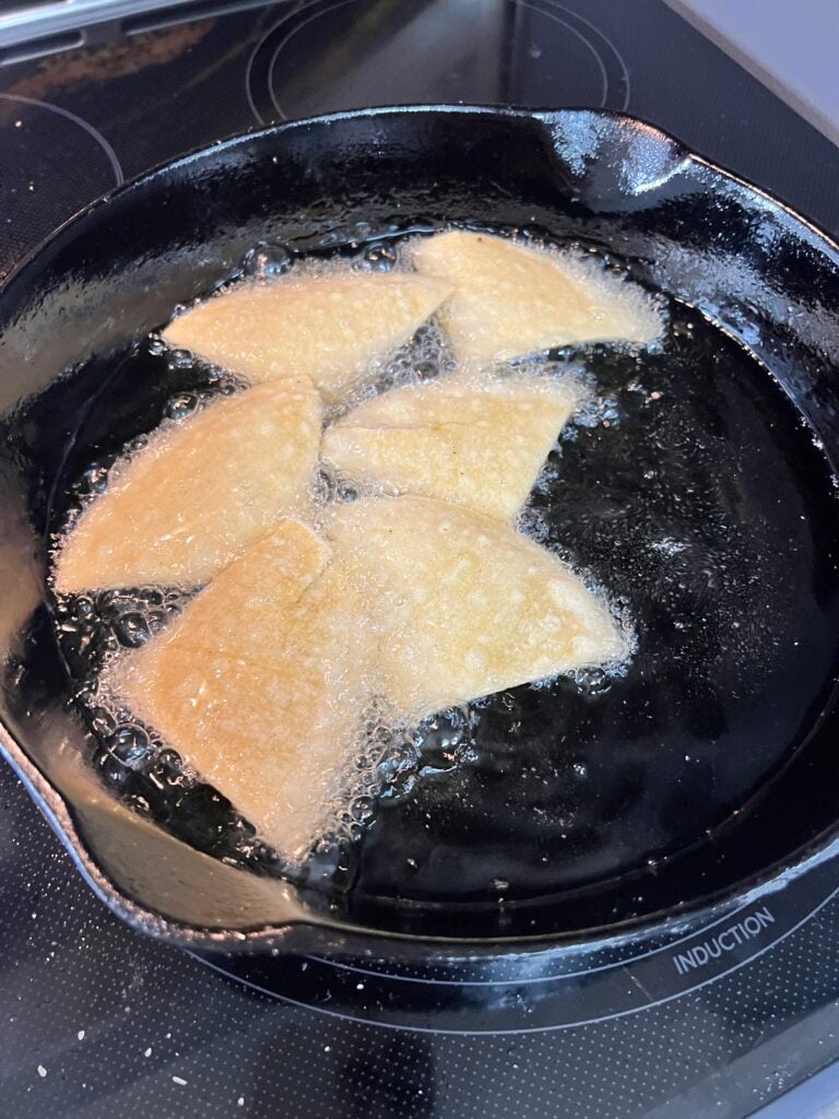 Corn tortilla triangles frying in hot 350F oil in a black cast-iron skillet, golden and crispy