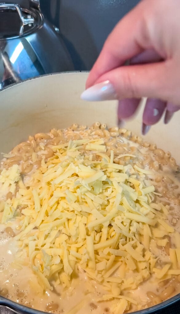 Shredded sharp cheddar cheese being stirred into creamy pasta with a white spoon