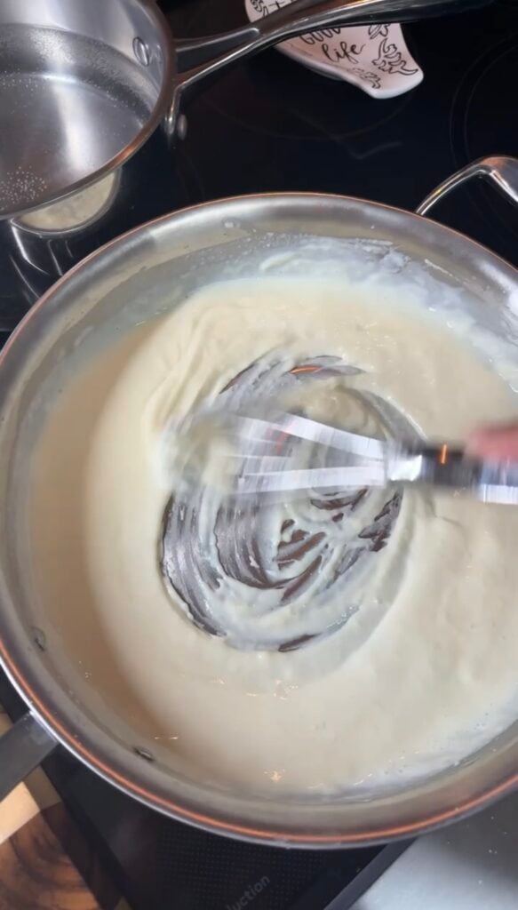 Active whisking of béchamel in an All-Clad pan, sauce still fluid as the milk begins to incorporate — motion blur on the whisk