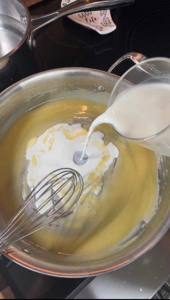 Cold milk being poured from an All-Clad measuring cup into a roux in a stainless pan, whisk ready to incorporate