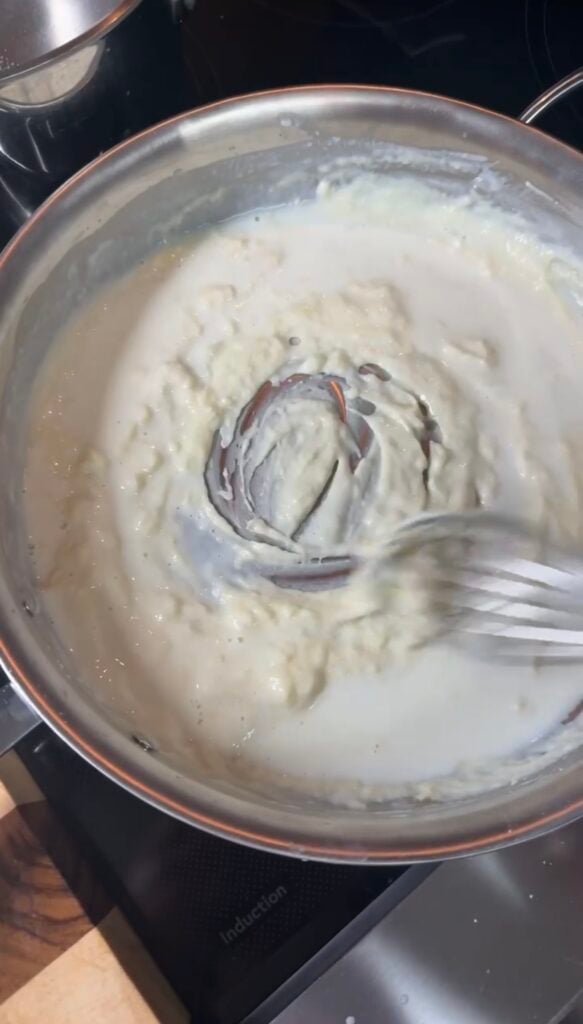 Béchamel sauce thickening in a stainless All-Clad pan, whisk pulled through leaving visible trails in the creamy sauce