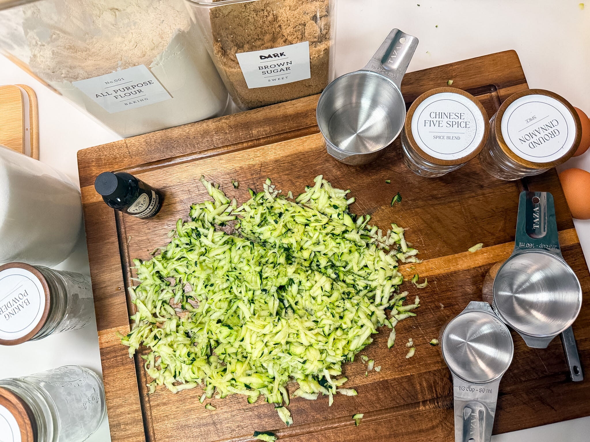 Shredded zucchini on a wooden cutting board surrounded by measuring cups with cinnamon, Chinese 5-spice, brown sugar, and other spices in small labeled glass jars