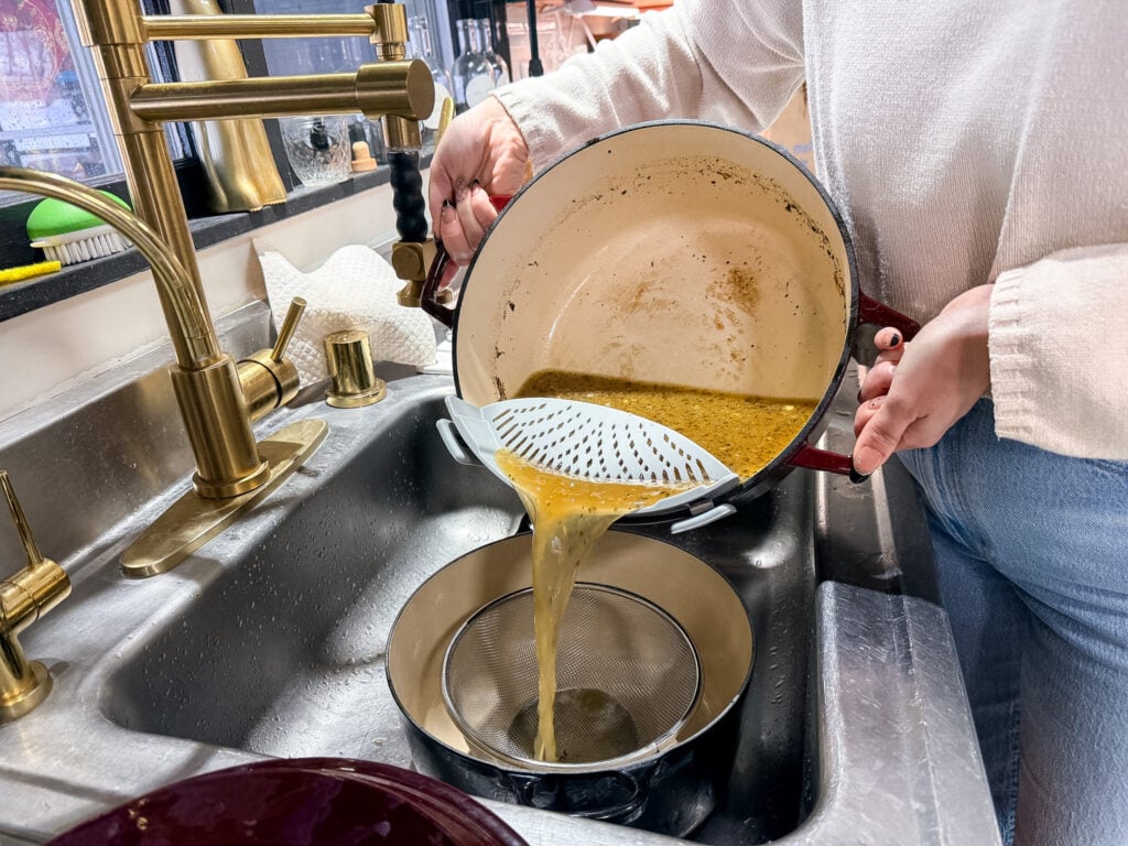 Golden broth streaming through a fine-mesh sieve into a stainless steel bowl below, showing the clarity and golden color of the finished broth during the straining process
