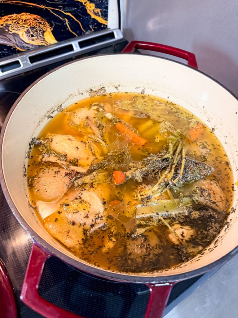 Golden chicken broth simmering gently in a cream-colored Dutch oven with bone-in chicken thighs, sliced carrots, celery, and aromatics visible in the pot