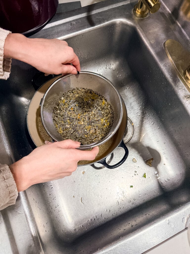 Fine-mesh sieve filled with strained solids from broth—cooked chicken pieces, carrots, celery, and herbs sitting in a stainless steel sieve over a sink